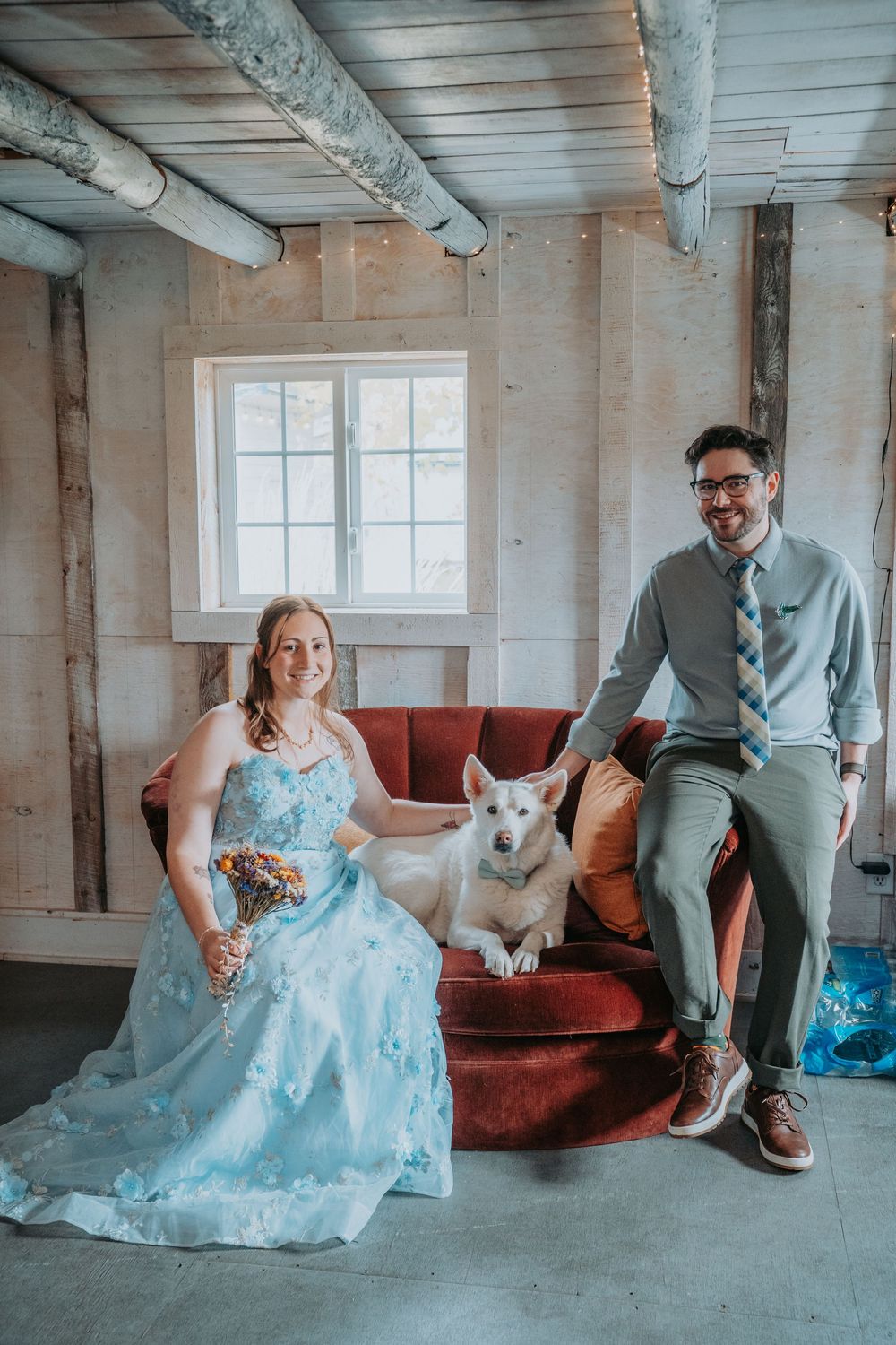 A couple in formal attire sits on a red couch with their white dog in an industrial-style room with white walls.