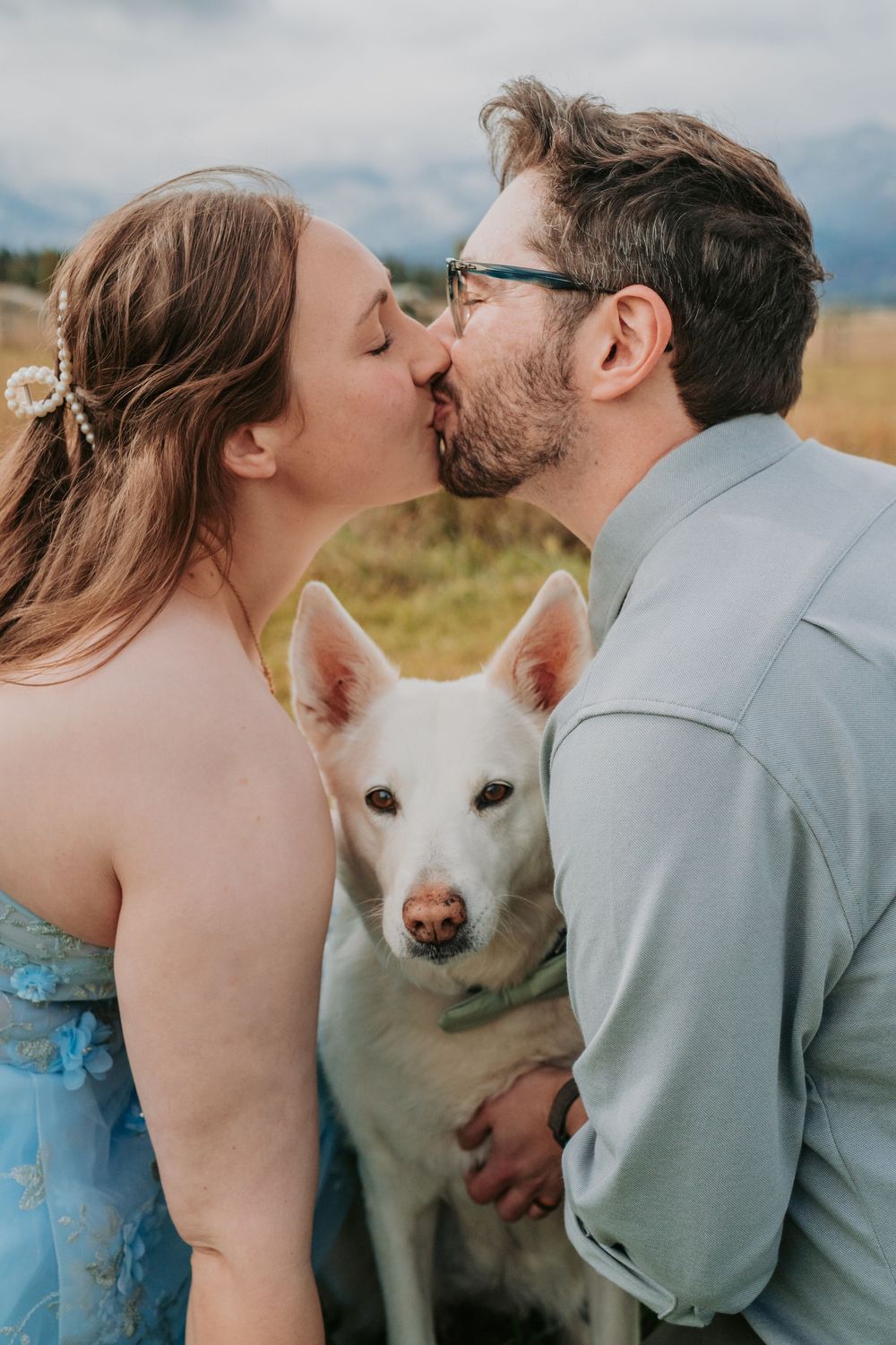 A couple shares romantic moments outdoors with a white dog photobombing their intimate poses against a mountain backdrop.