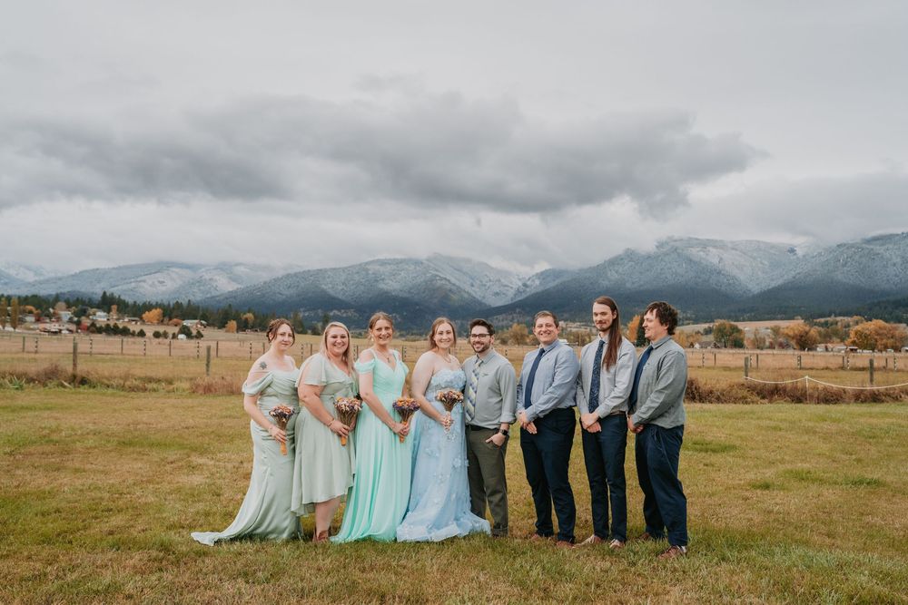 Wedding party in formal attire poses together against scenic mountain backdrop with cloudy skies.