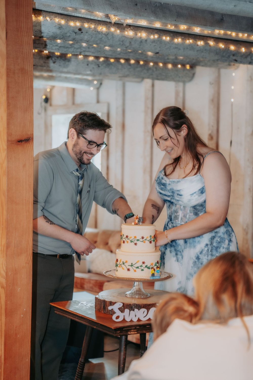 A couple cuts their wedding cake together at a rustic venue decorated with string lights.
