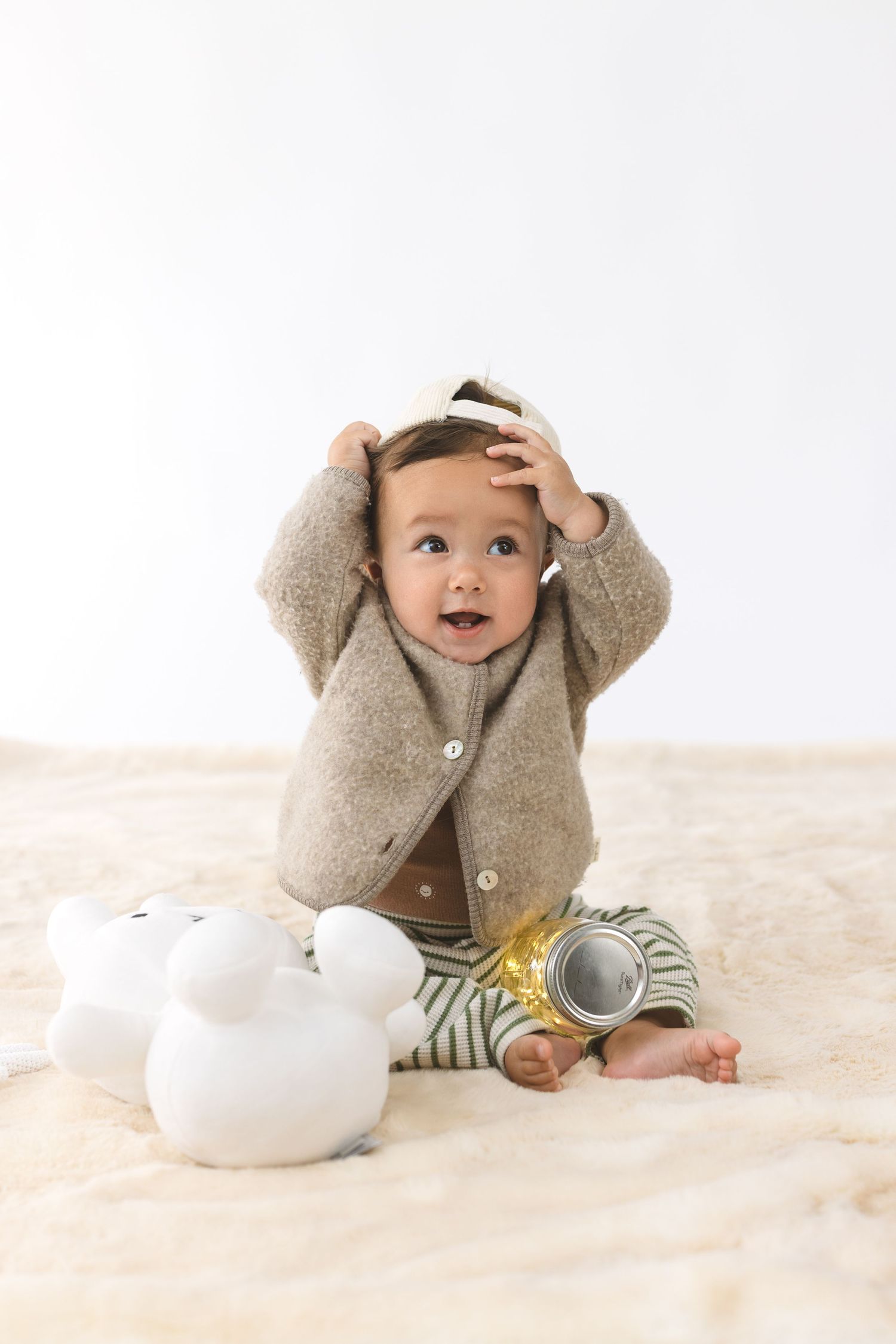 Baby in cozy gray cardigan sweater waves while sitting against white background in lifestyle portrait series.