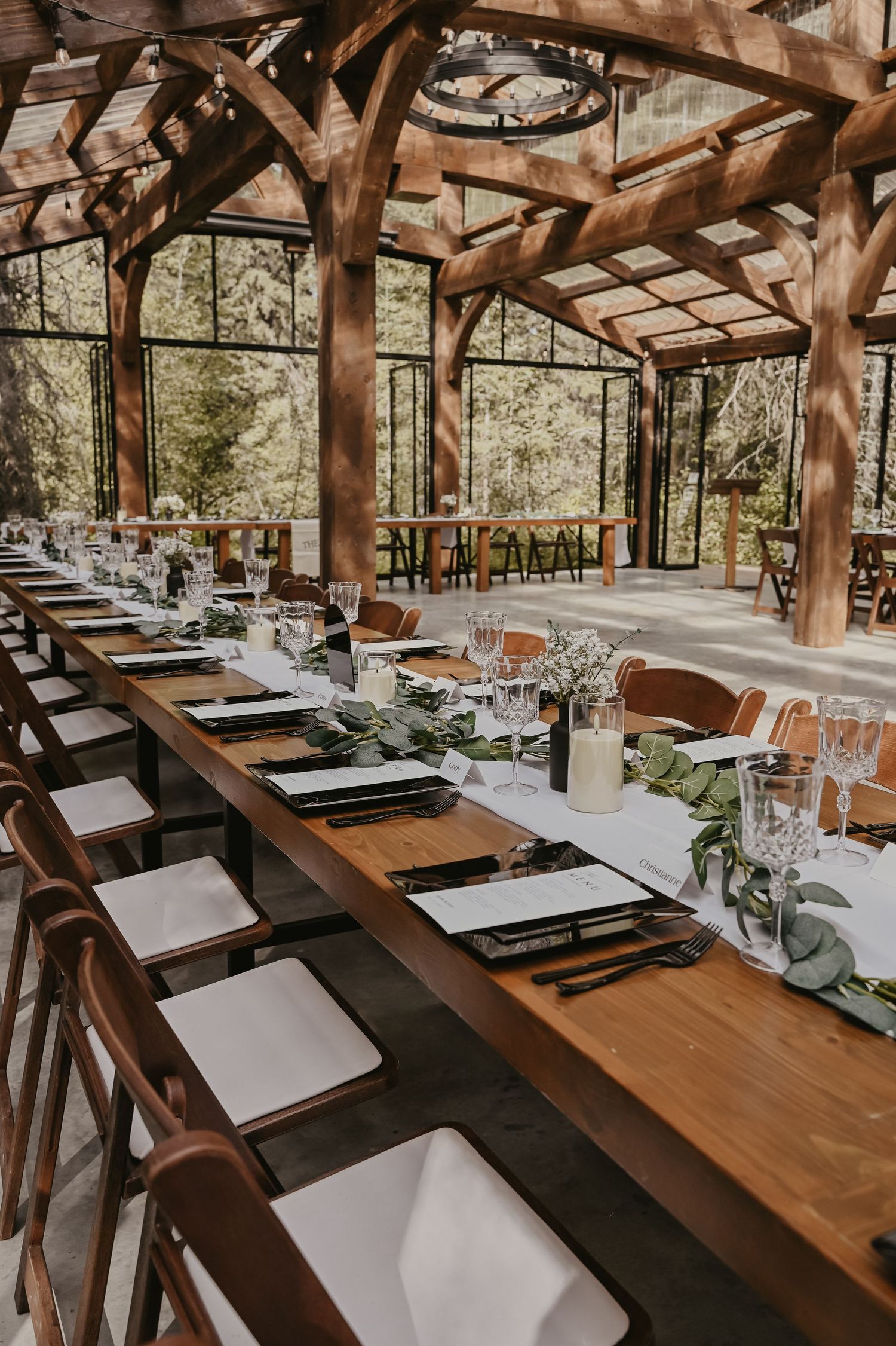 Elegant wedding reception table setting in rustic wooden pavilion with glass windows and natural light.