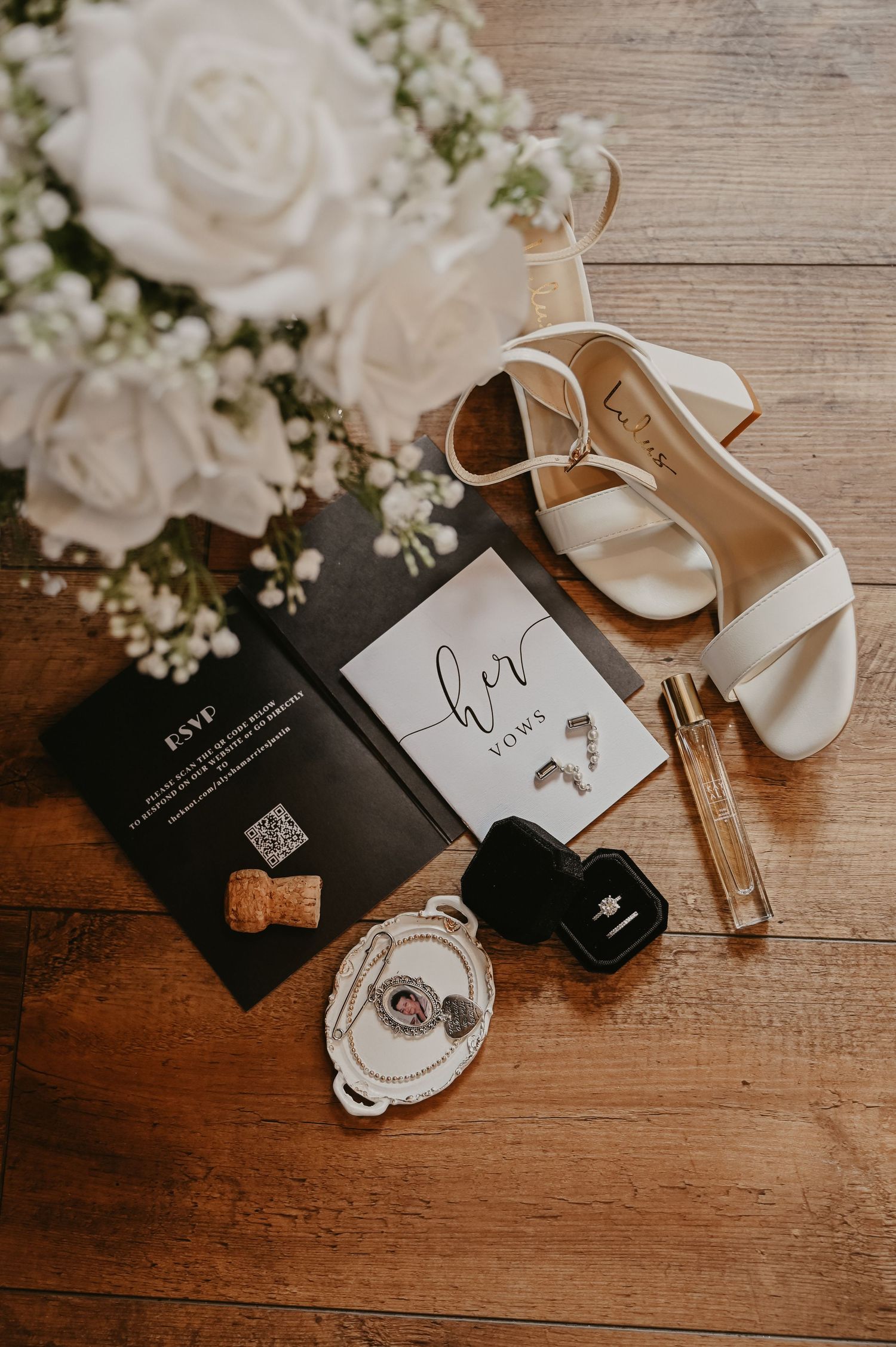 Wedding details including white roses, invitation cards and bridal shoes arranged on wooden surface.
