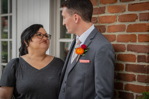 A couple shares an intimate moment against a brick wall backdrop, with one wearing a gray suit and bright boutonniere.