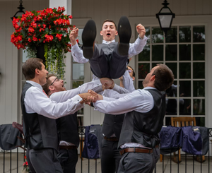A group of groomsmen in gray vests playfully celebrate during a wedding photo session in front of a building.