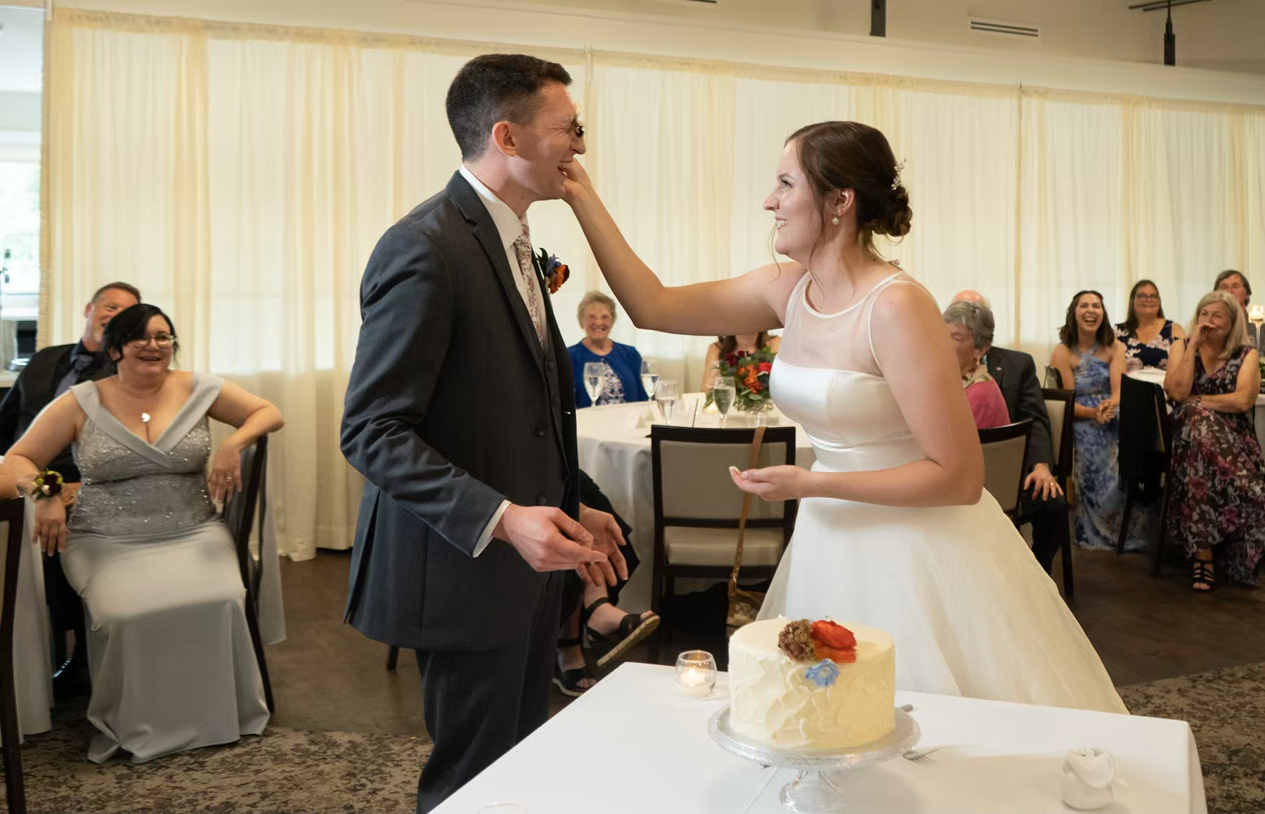 A couple shares a sweet moment while cutting their wedding cake at a reception venue with white tablecloths.