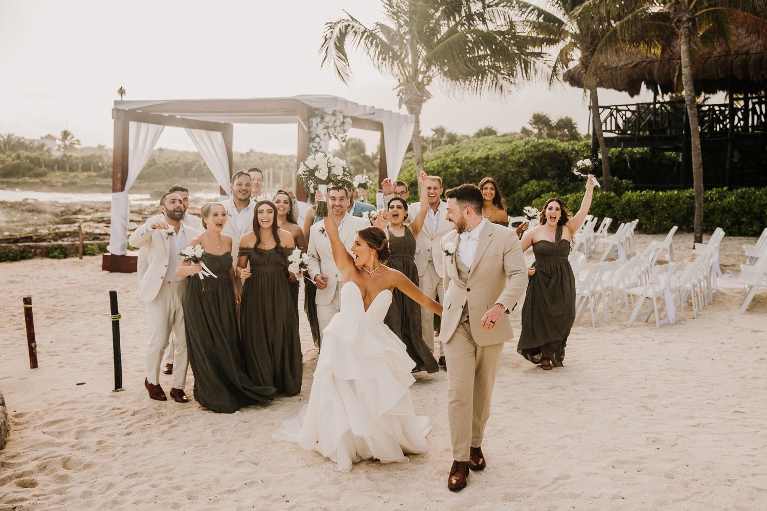 A wedding party celebrates joyfully on a tropical beach with a white canopy and palm trees in the background. El Dorado Royale Wedding Photo