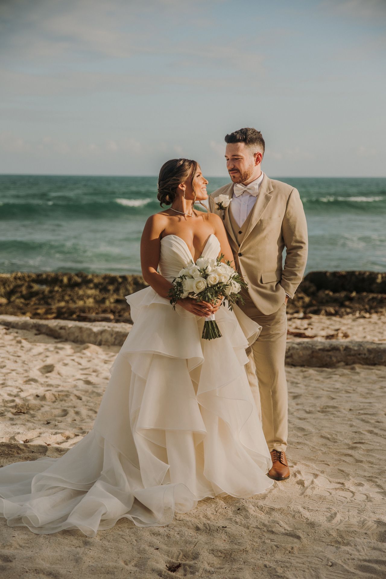 A couple shares romantic moments on a scenic beach with waves and rocks in the background during their wedding photoshoot. El Dorado Royale