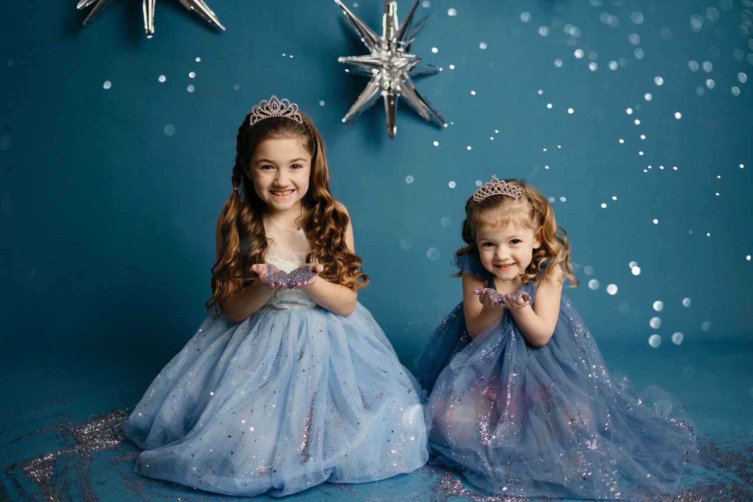 Two children in sparkly princess dresses pose together against a starry blue backdrop with twinkling lights.