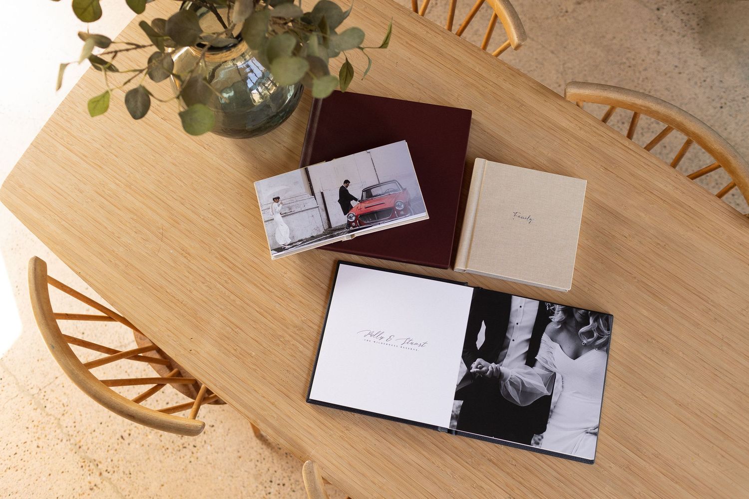 Photo albums and cards arranged on a light wooden table with decorative chair and small potted plant visible.