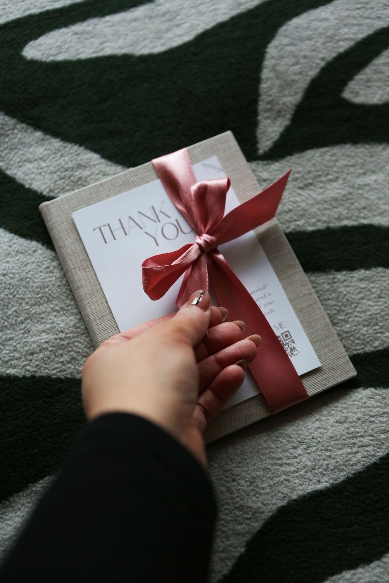 A hand holds a white 'Thank You' card decorated with a pink satin bow against a dark patterned carpet background.