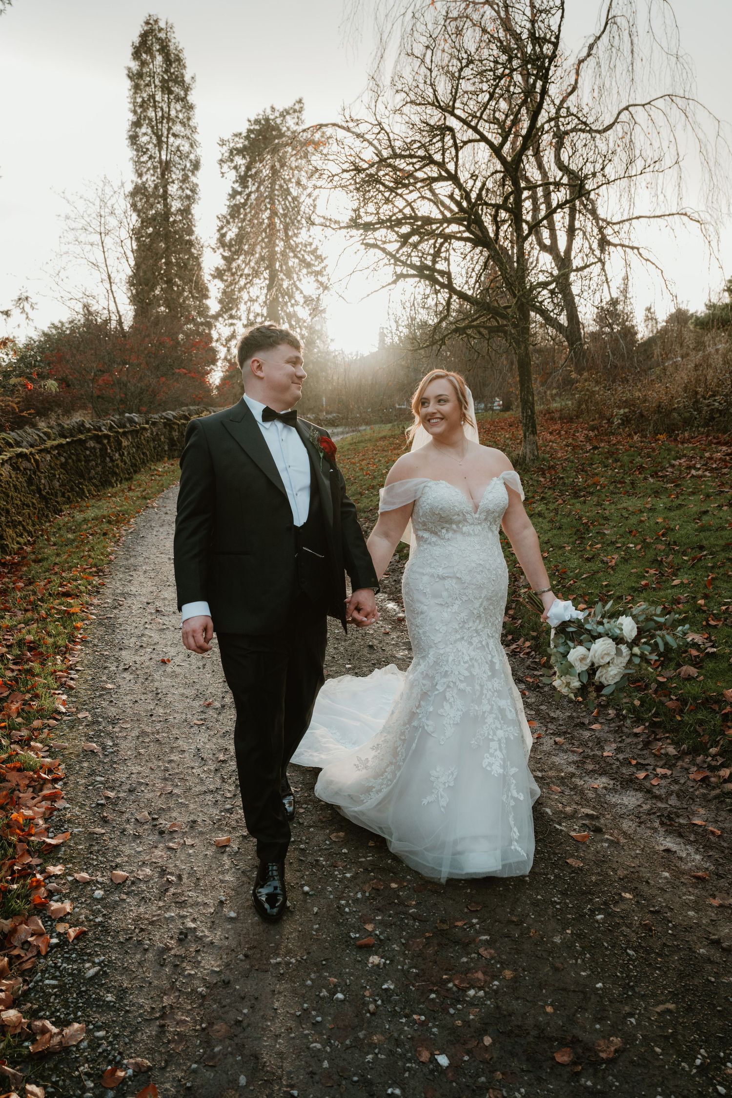 A couple in wedding attire walks along a scenic autumn path during golden hour as fallen leaves cover the ground.