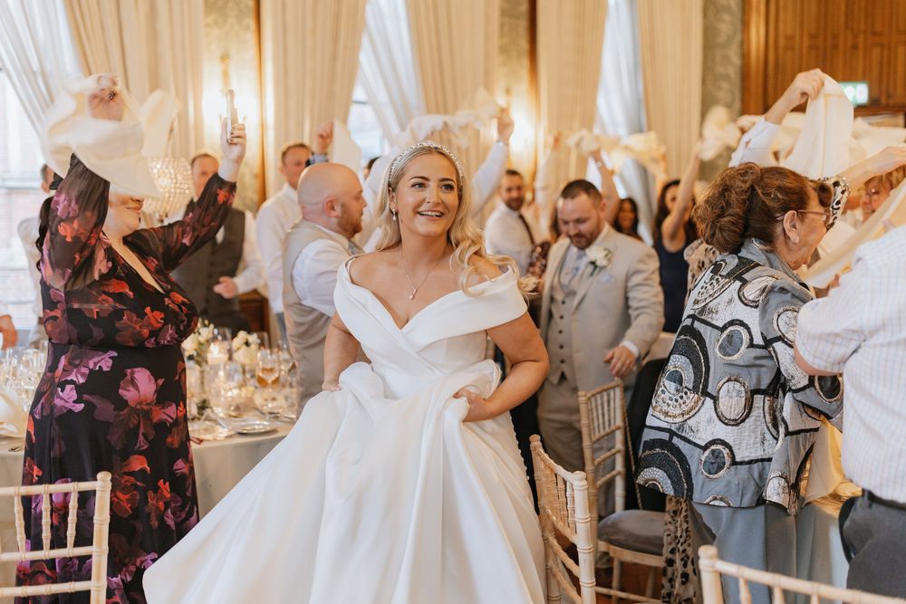 Wedding reception guests dancing and celebrating with joyful bride in elegant white off-shoulder gown at elegant venue.