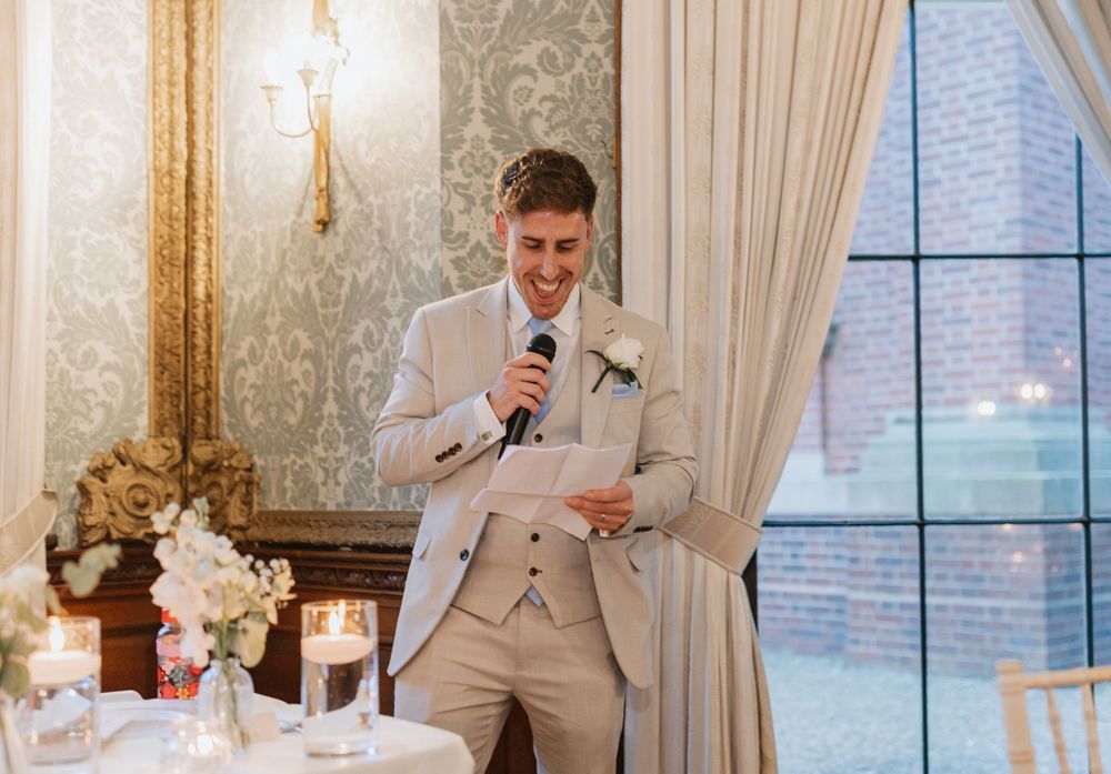 Wedding party members in beige suits pose for photos in an elegant ballroom with white curtains and chandeliers.