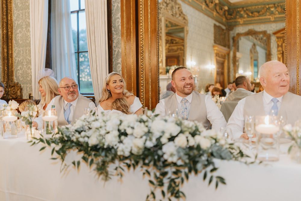 Elegant wedding reception table settings with white floral centerpieces and greenery in an ornate ballroom with chandeliers.