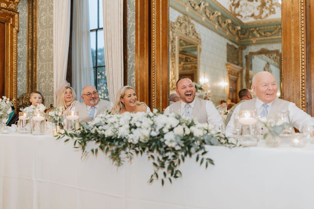 Elegant wedding reception in an ornate ballroom with white floral centerpieces and gilded mirrors on the walls.