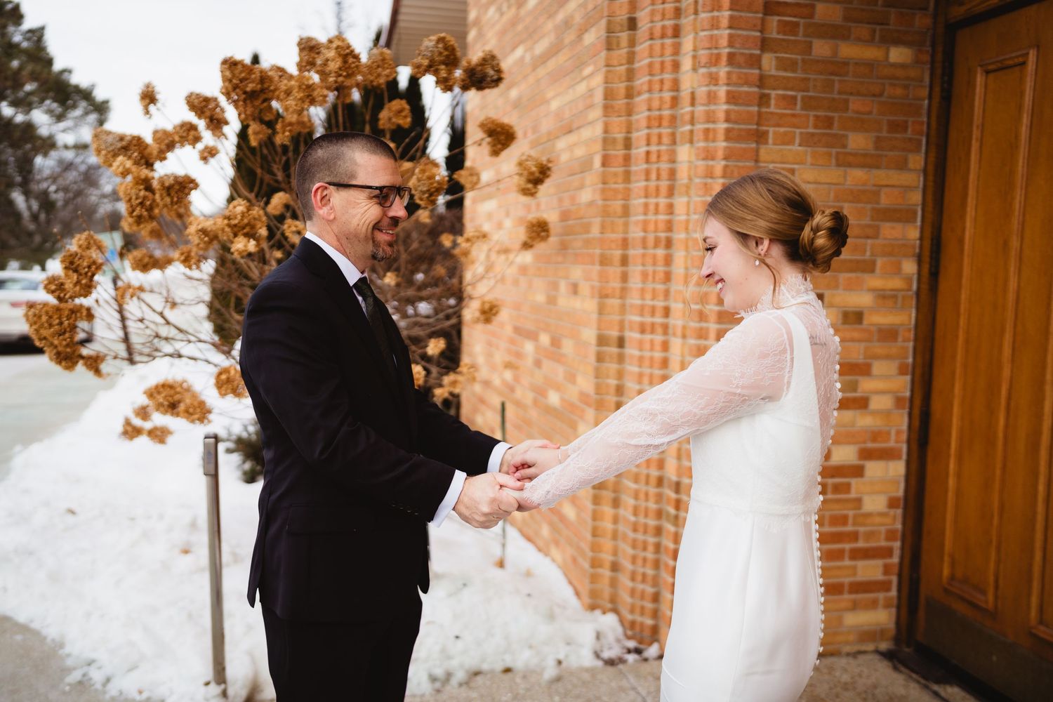 A romantic first look moment between newlyweds against a brick wall backdrop with snow-covered trees.