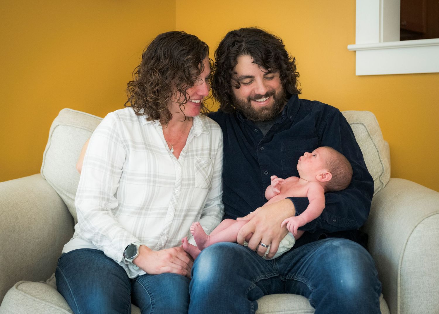 A couple sits together on a beige couch against a yellow wall, holding a newborn baby and sharing a tender moment.