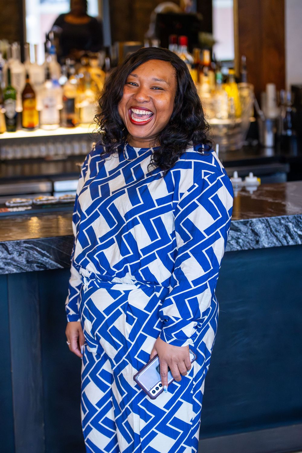 Woman wearing blue and white geometric print dress laughs joyfully at bar with bottles in background.