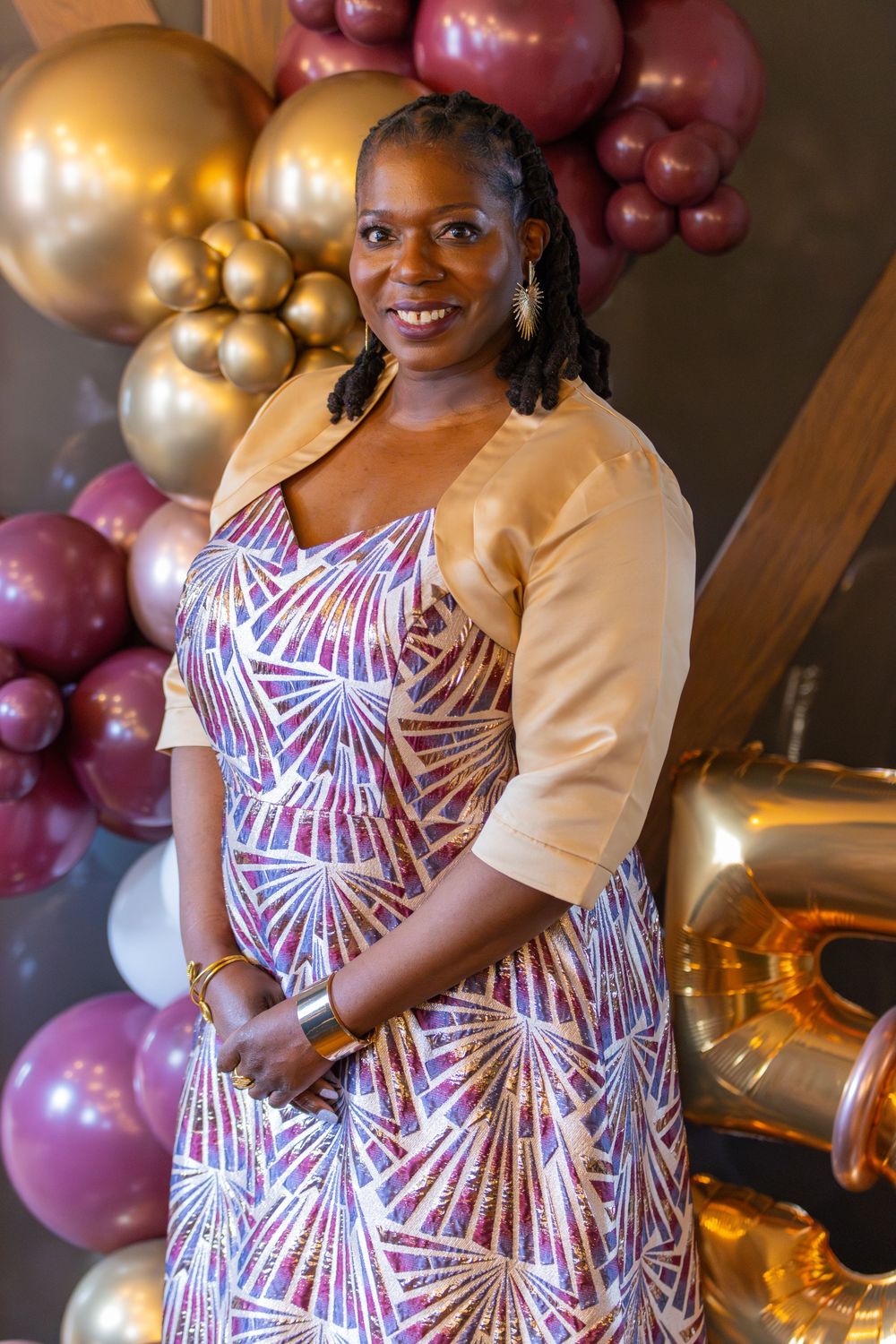 Woman in patterned dress poses in front of festive gold and pink balloon display at celebration.