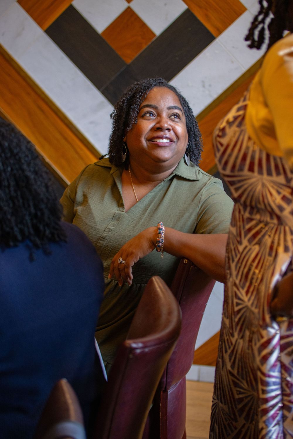 A person in an olive green top sits in front of a decorative wall with a warm, engaging expression.