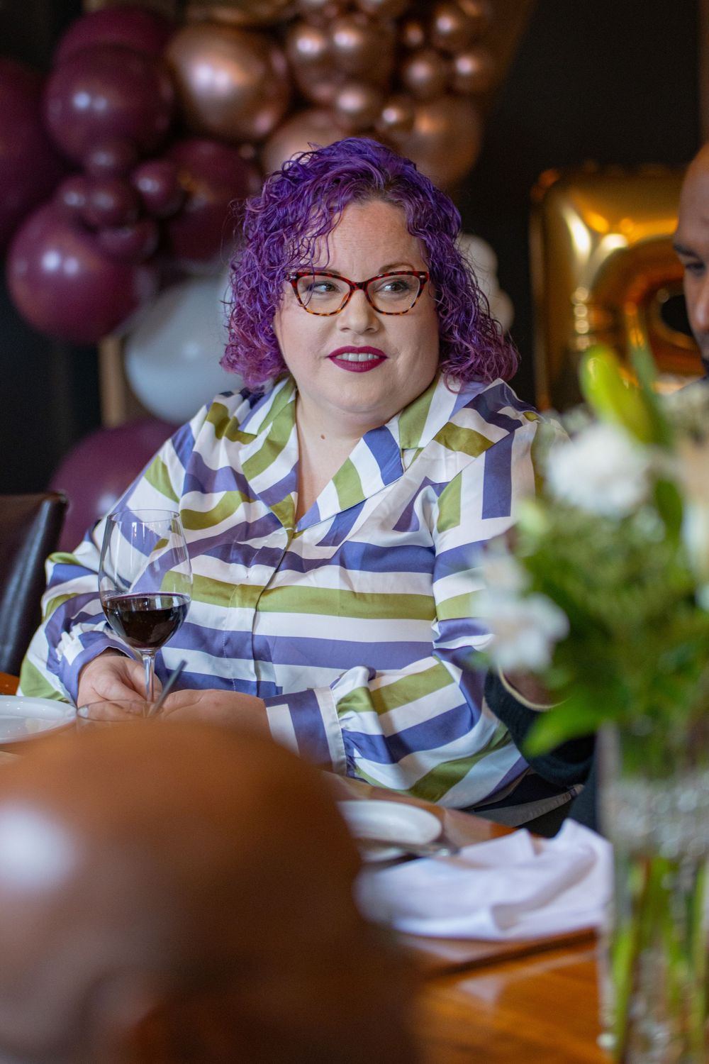 Person with purple hair and glasses enjoying wine at an elegant dinner table setting.
