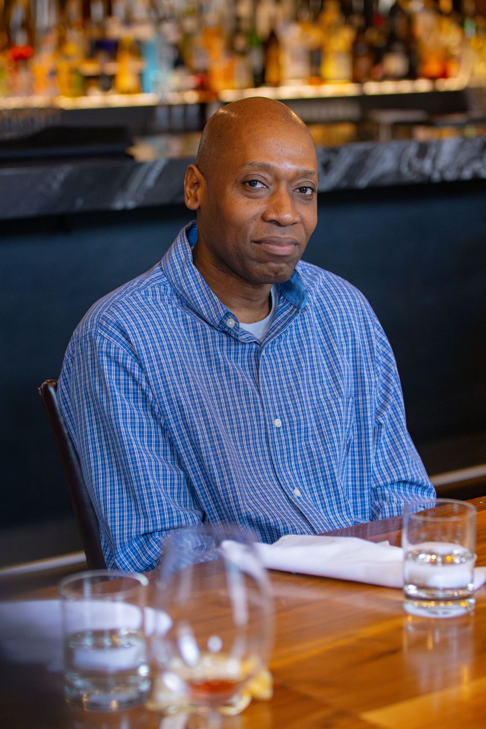 Man in blue checkered shirt sitting at wooden bar table with drinks.