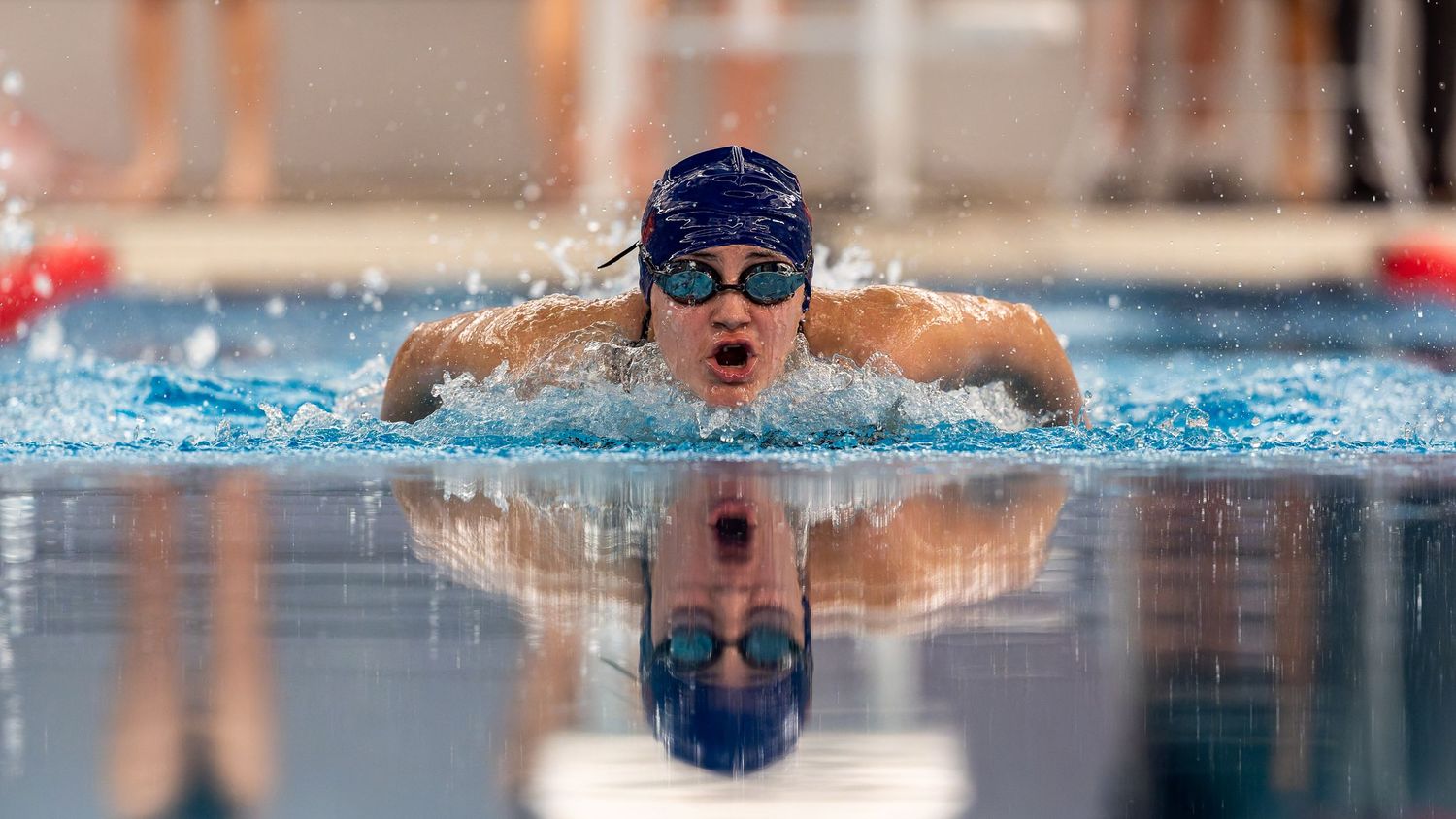 Competitive swimmer performs butterfly stroke in pool with perfect reflection in water during race.