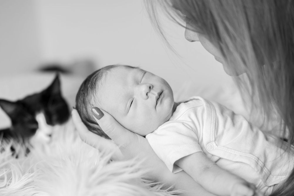 Sleeping newborn baby cradled by mother with a cat in the background, black and white photo.