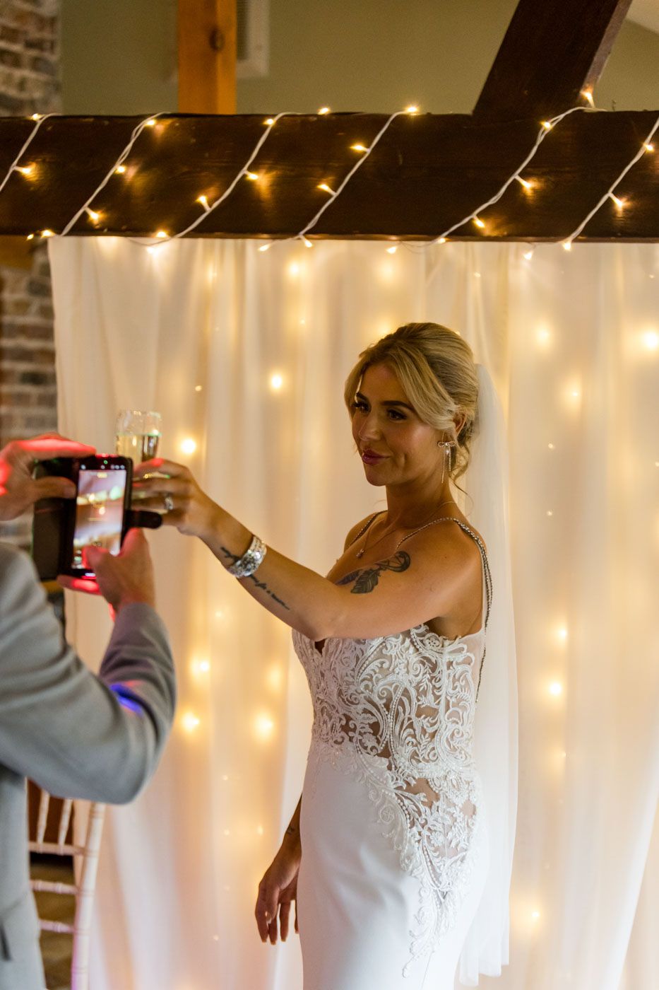 A bride in a white lace wedding dress poses against a backdrop of twinkling string lights.