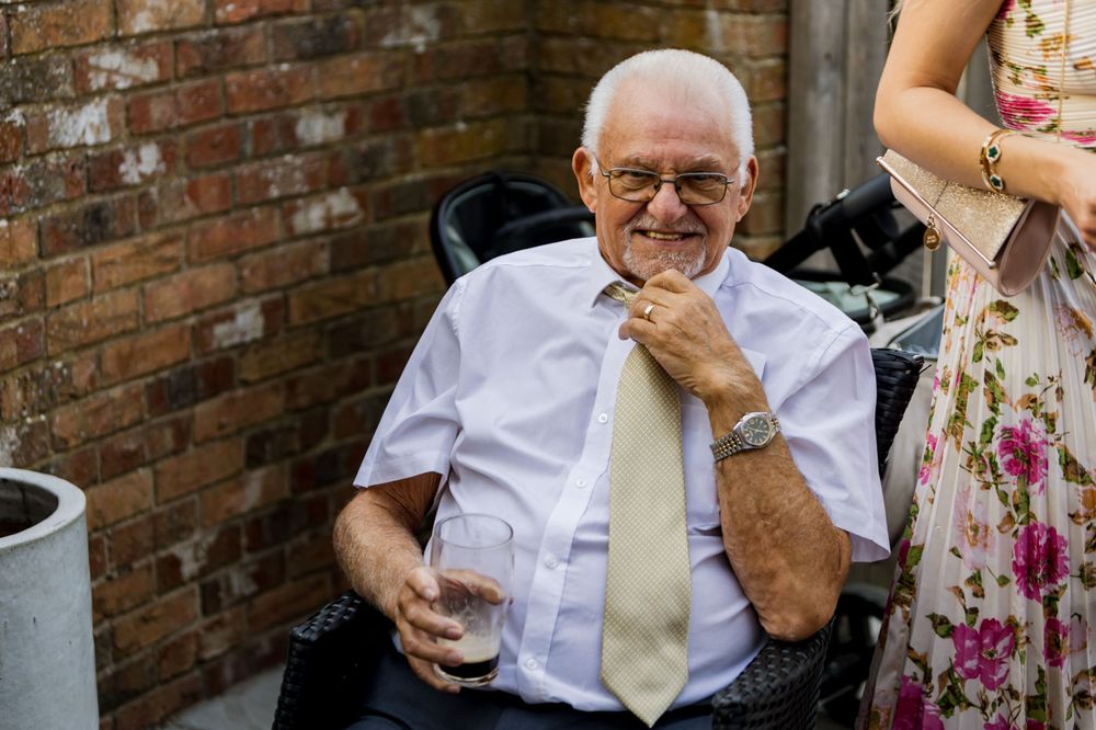 An elderly gentleman in a white shirt and tie smiles warmly while seated outdoors by a brick wall.