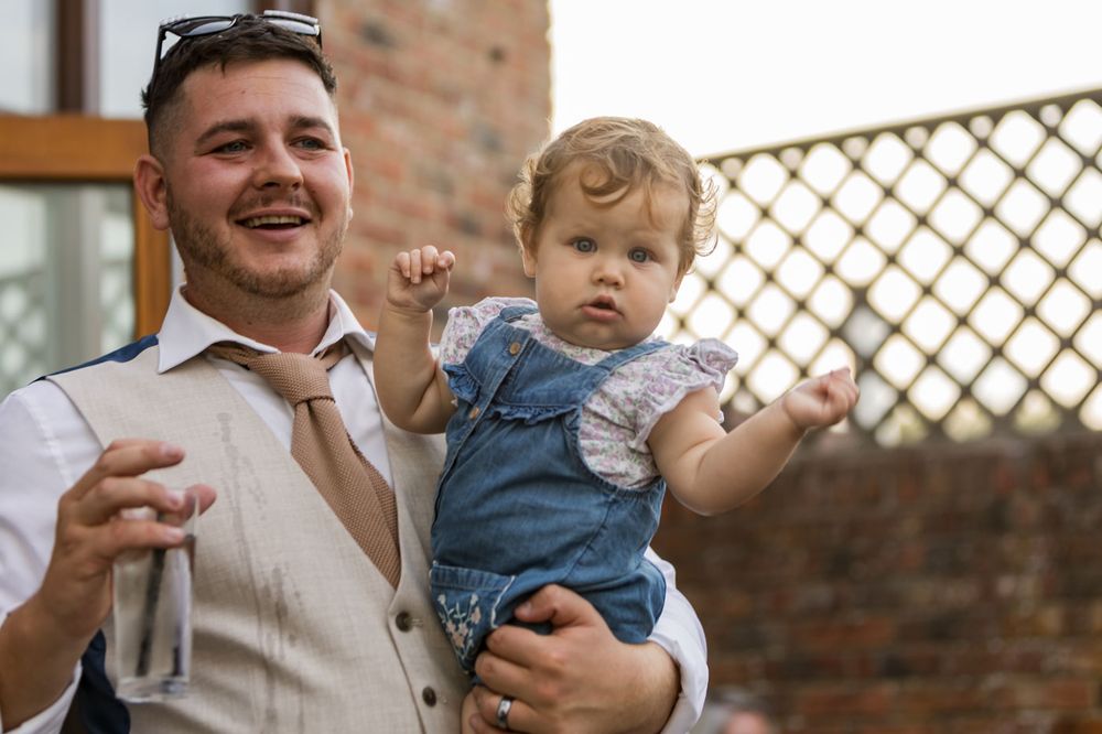 A toddler in denim overalls is held during an outdoor celebration against a brick wall and lattice backdrop.