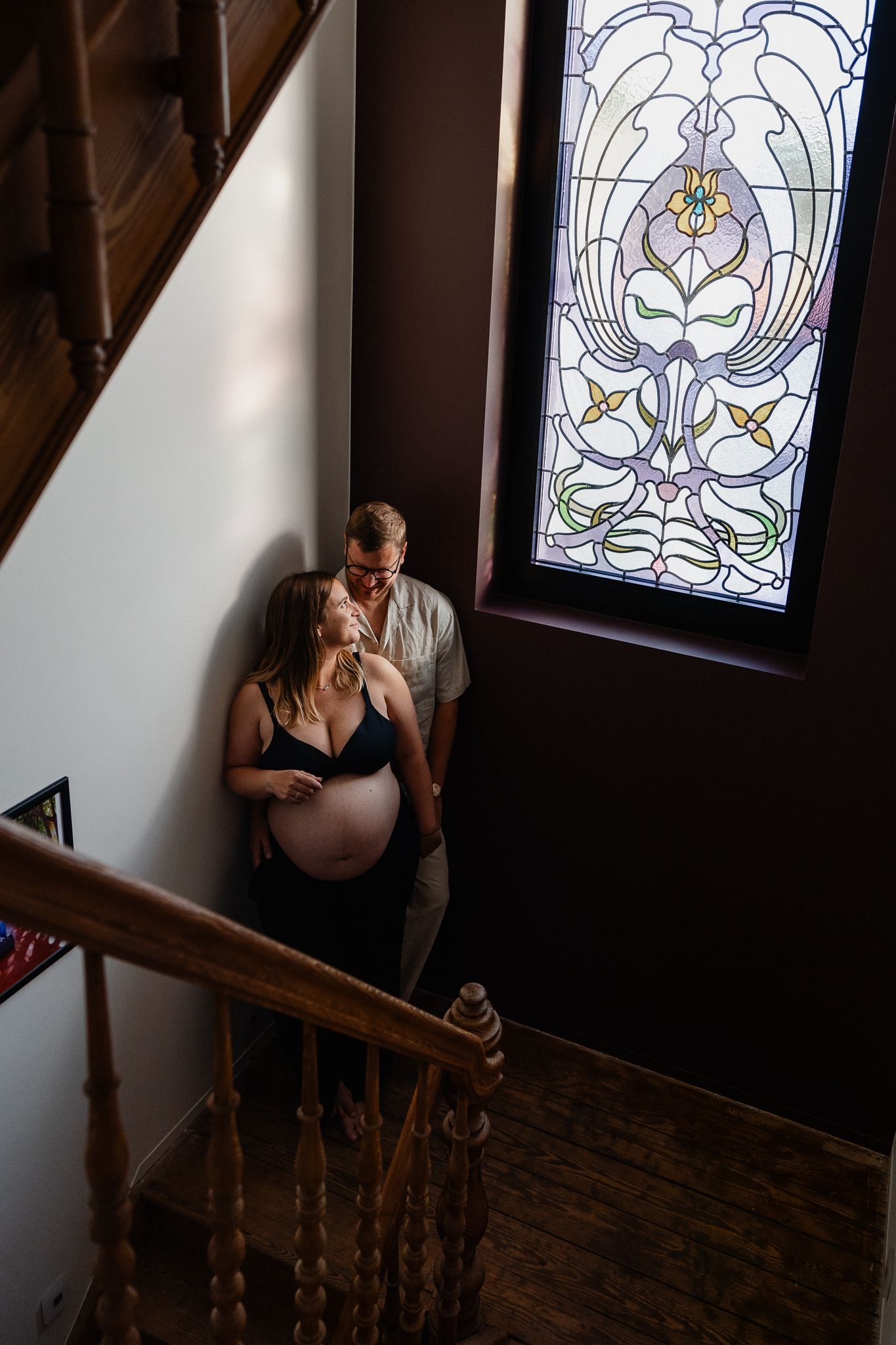 Femme enceinte et futur papa dans l’escalier de leur maison, lumiere coloree filtrant a travers un vitrail art deco, seance photo grossesse