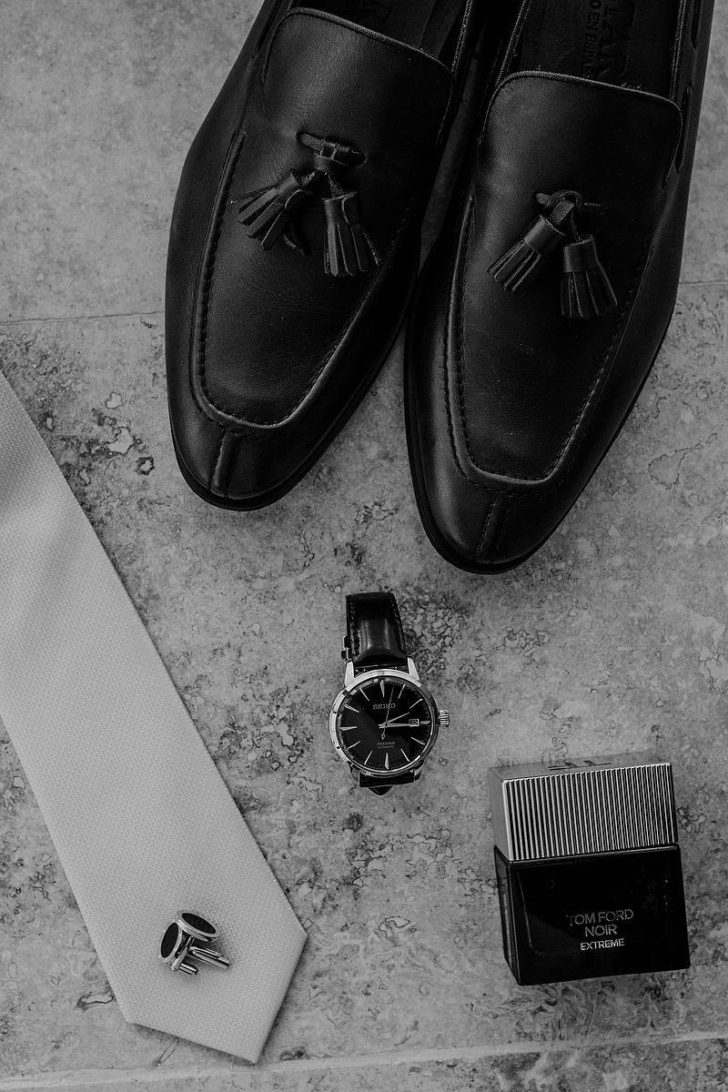 Black leather loafers arranged with a watch and comb on a textured gray surface.