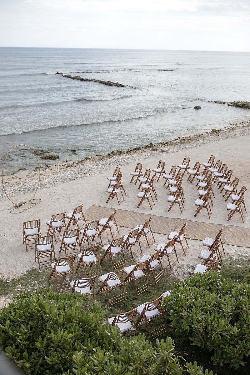 Empty white beach chairs arranged in rows on a sandy shoreline with green shrubs and gentle waves in the background.