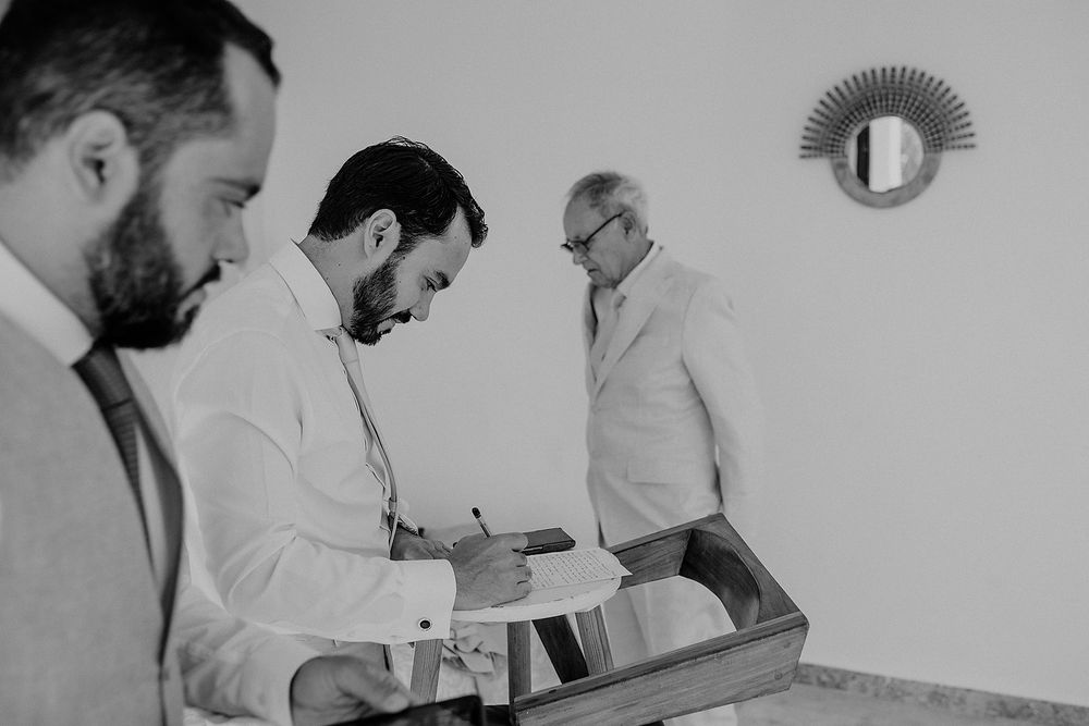 Three men in formal attire review documents together in a minimalist room with a sunburst mirror.