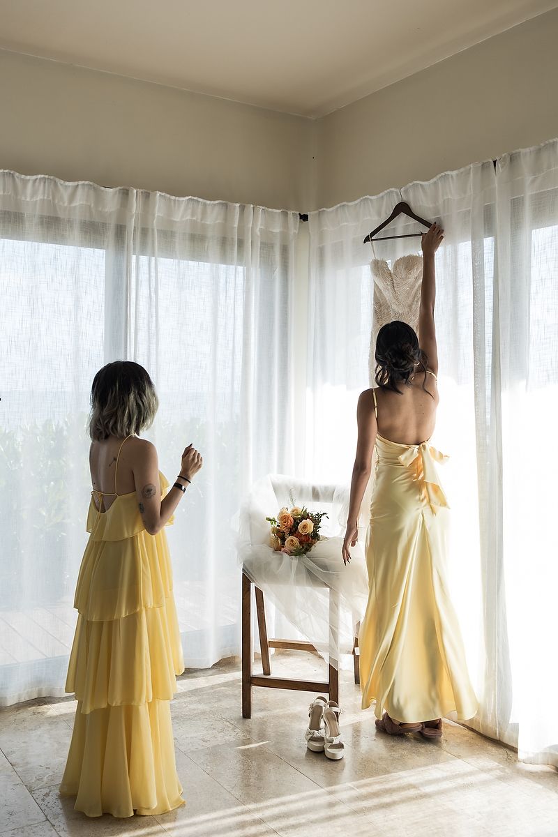 Two women in yellow dresses prepare for a wedding, with a dress hanging by sheer curtains.