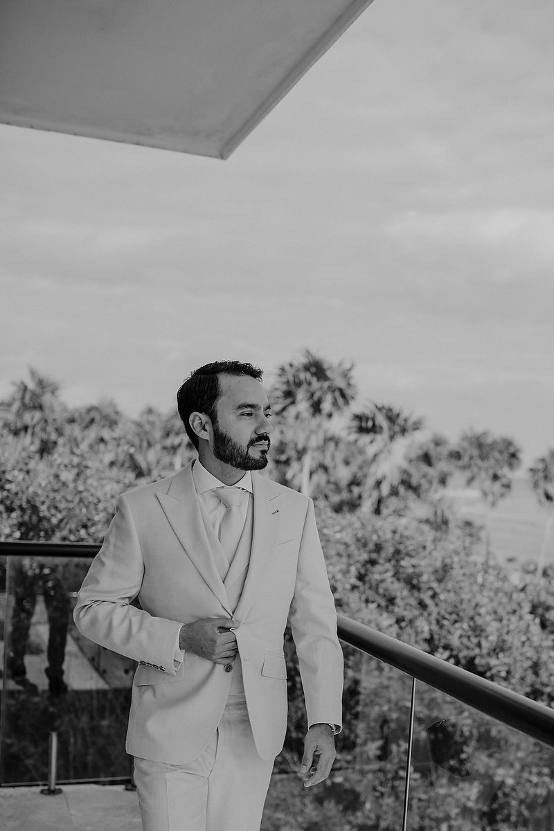 A person in a white suit stands on an outdoor deck with greenery visible in the background.