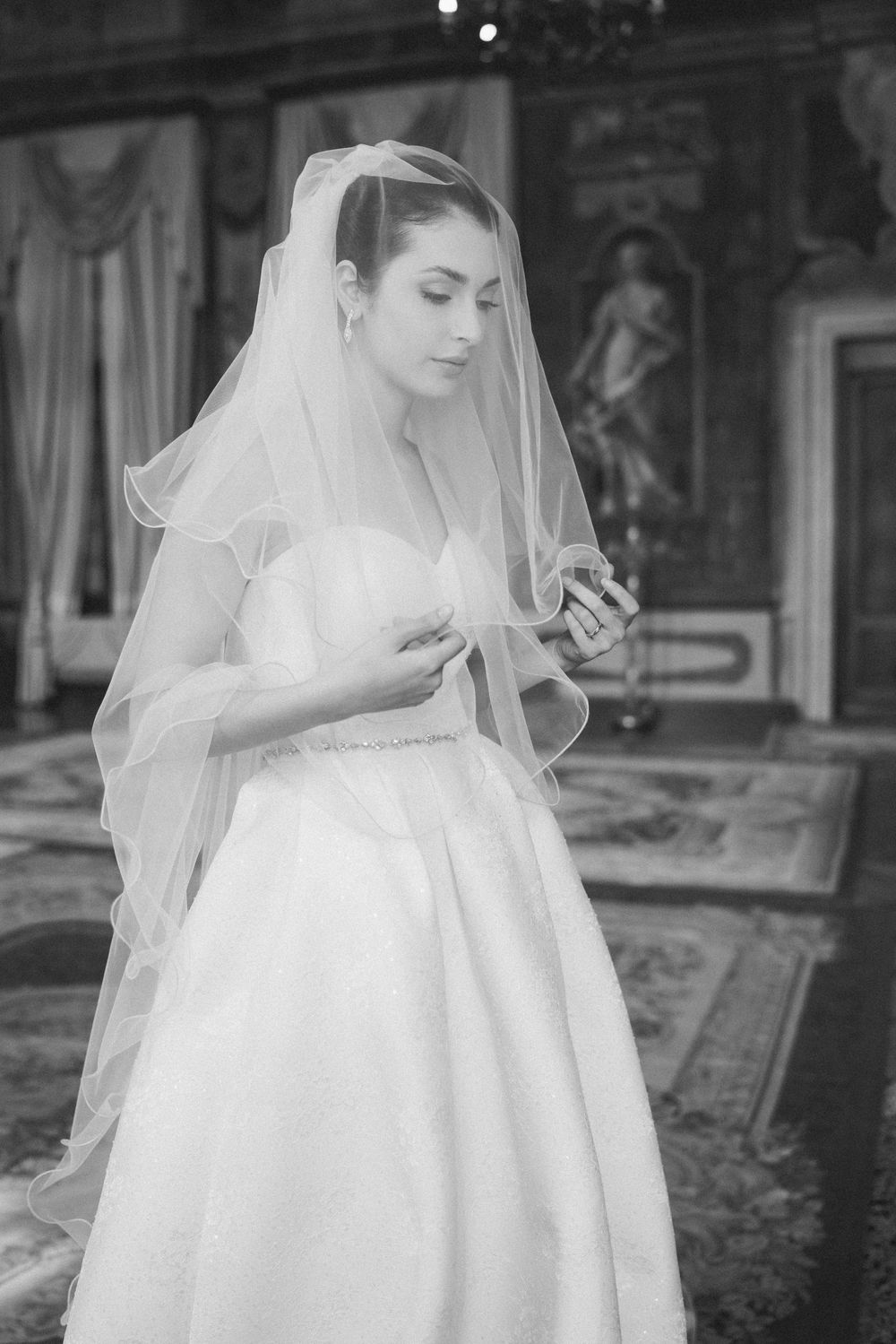 A bride in an elegant white wedding gown and veil stands in an ornate room looking down at something in her hands.