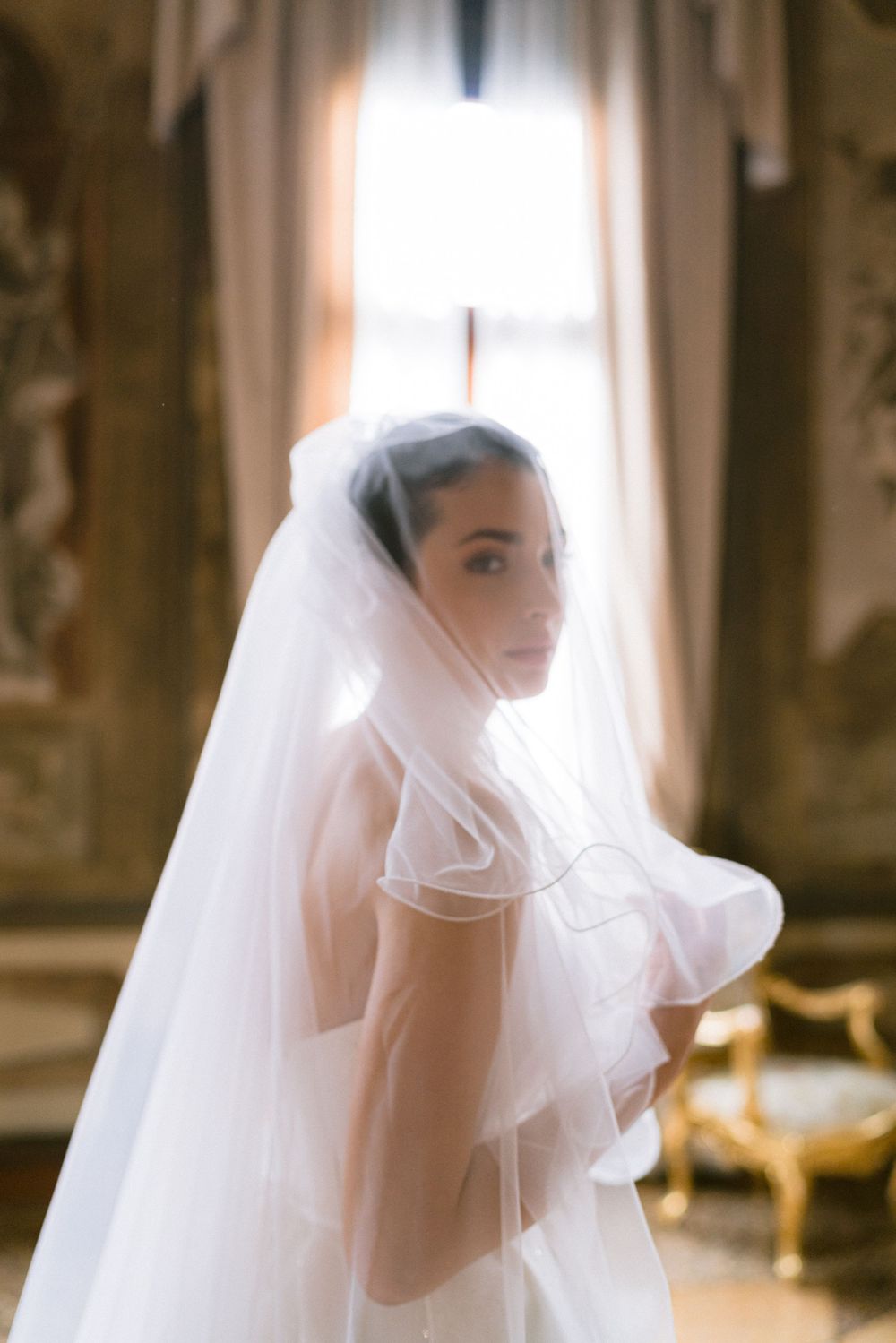A bride in a flowing white veil is captured in a series of ethereal portraits with soft window lighting. Wedding photoshoot in Venice