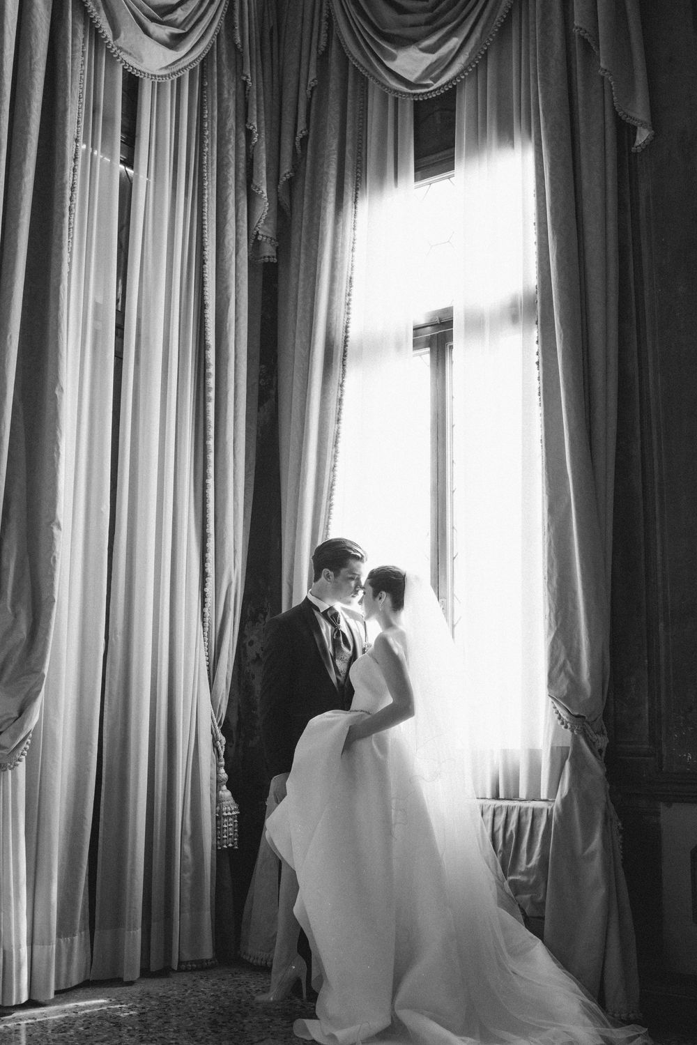A black and white photo series of a bride in a flowing wedding dress standing near elegant curtained windows.