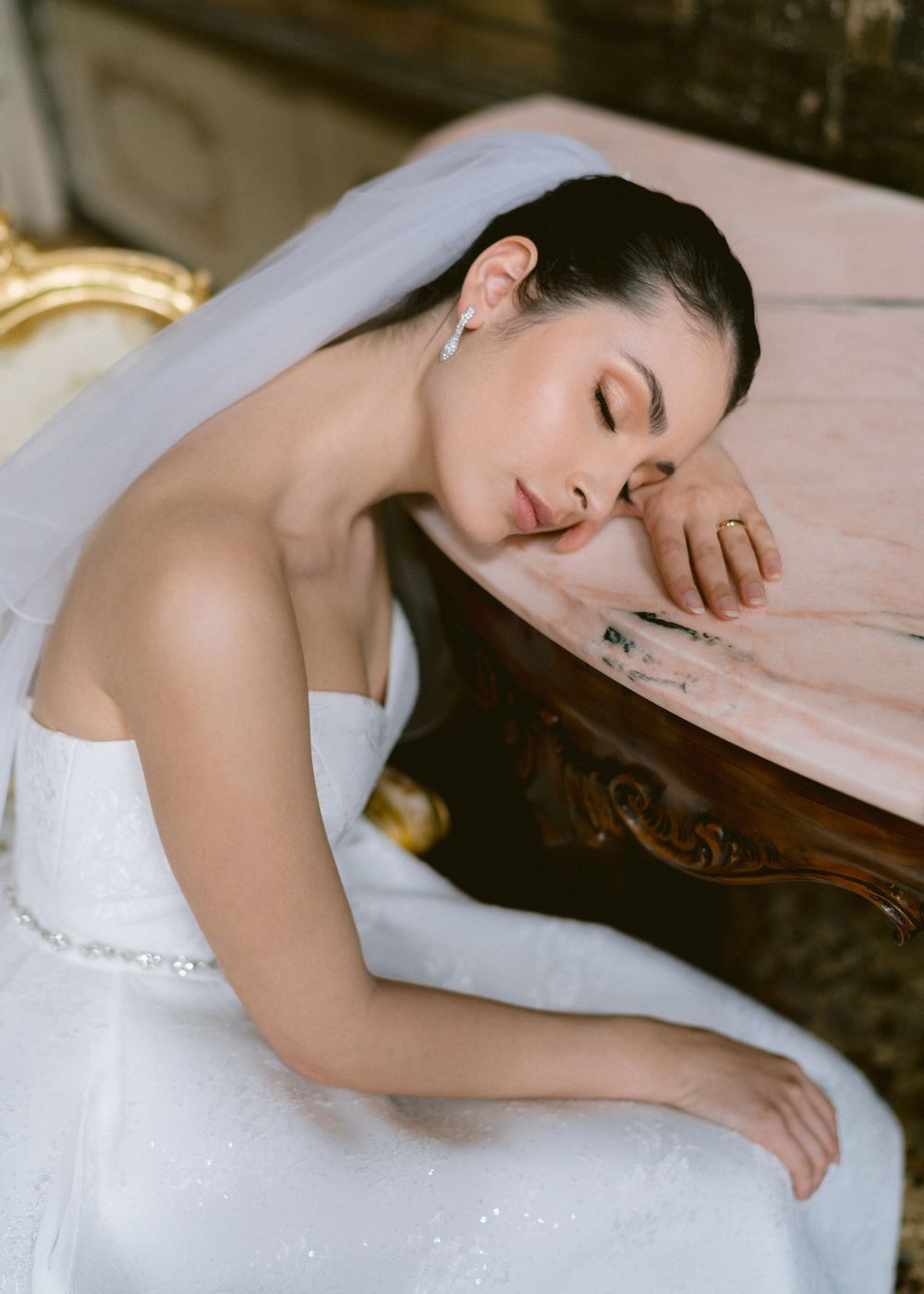 A bride in a white wedding dress lies gracefully on a pink chaise lounge in an elegant setting.