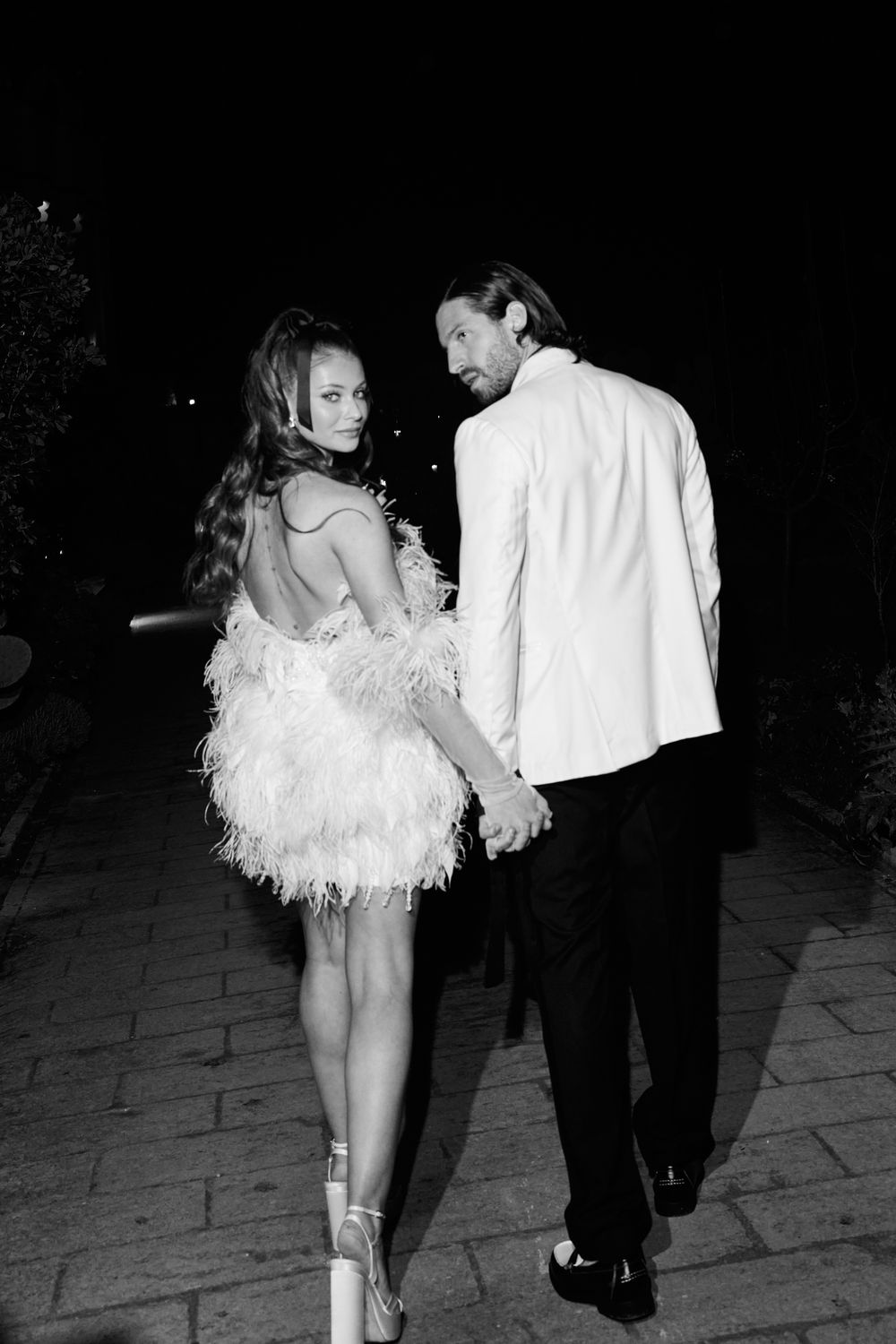 A couple in formal attire walks away on a dark path, with the person wearing a white feathered dress. Puglia wedding photographer