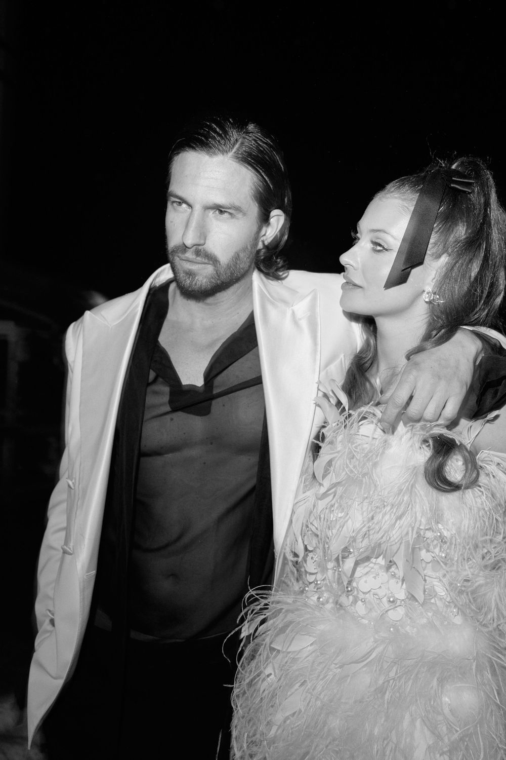 A black and white photo of a couple posing together, with the woman wearing a feathered dress and the man in a white blazer.