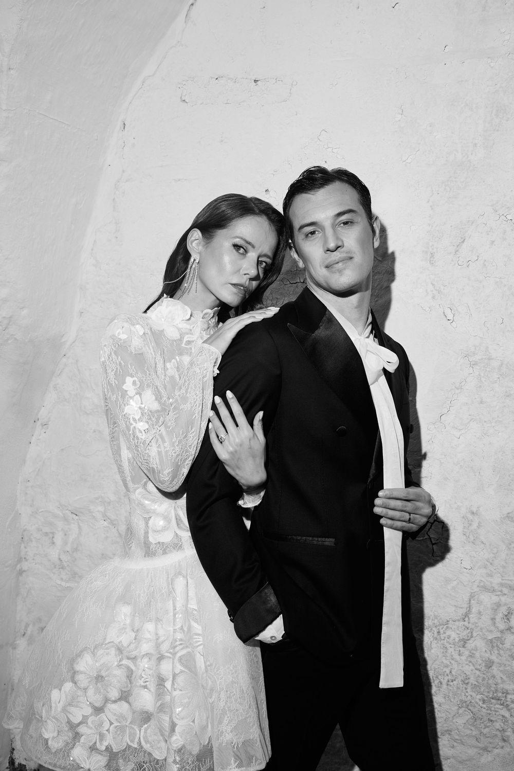 A couple in formal black attire poses together against a white textured wall in a series of artistic black and white photos.