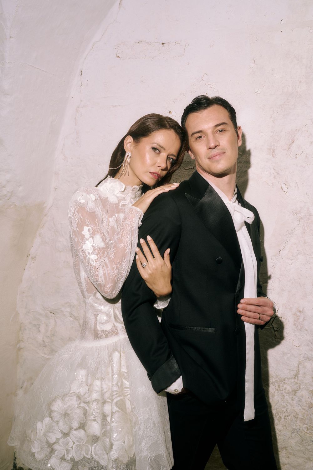 Elegant couple in wedding attire pose together against textured wall in romantic bridal portrait.