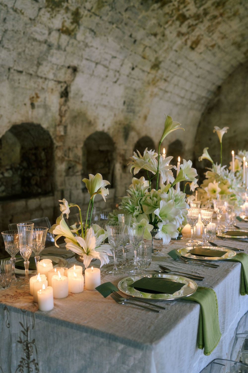 An elegant candlelit dinner table set in a stone cellar with white lilies and glowing votives along the length.
