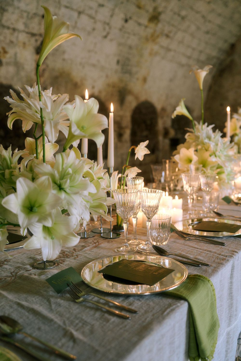 Elegant table setting with white lilies, candles, and sage green accents on a textured linen tablecloth.