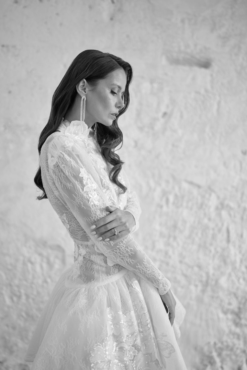 Elegant black and white portrait of a bride in a long-sleeved lace wedding gown with delicate details against a textured wall.