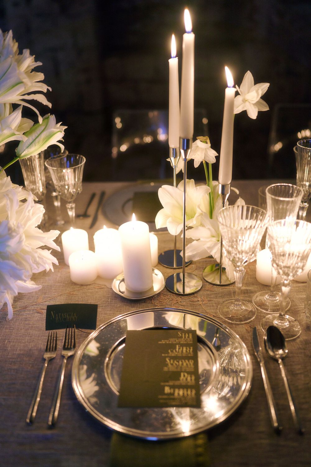 Elegant table setting with white candles, crystal glassware and white lilies creating a romantic evening ambiance.